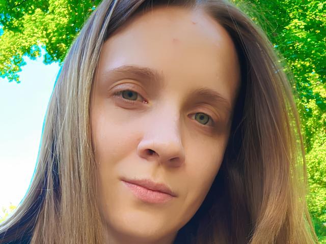 CristalLora69 tem olhos verde-claros, cabelo castanho liso e um sorriso discreto com lábios rosados. Seu nariz é pequeno e ela está olhando ligeiramente para a direita..