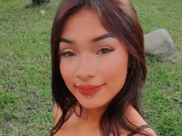 SashaHans has dark brown hair, a nose ring, and a lip piercing with a small ball. She's smiling with red lipstick, and has dark, well-defined eyebrows.