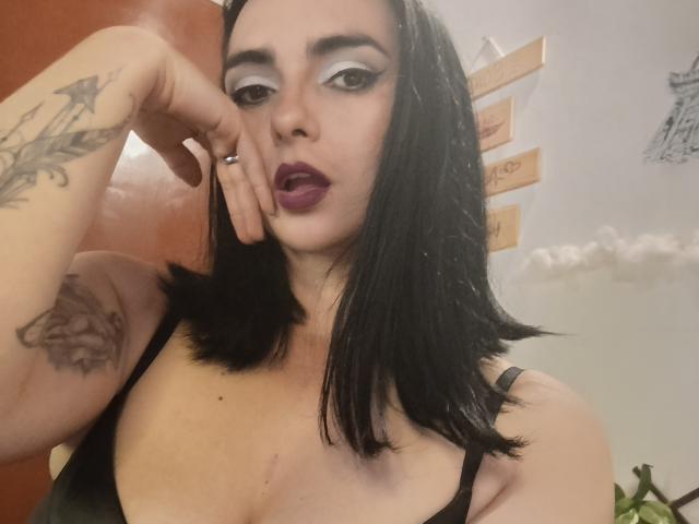CandyParis mit schwarzen, schulterlangen Haaren, trägt dunkles Make-up, einschließlich weißem Eyeliner und lila Lippenstift, ein schwarzes Tanktop und zeigt Tattoos am Arm. Ihre Hand ist in der Nähe des Mundes, und sie schaut in die Kamera..
