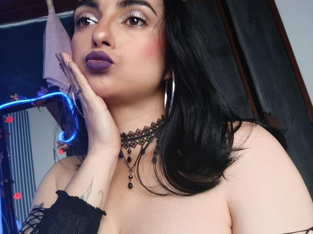 CandyParis with purple lipstick, black hair, hoop earrings, and a black choker, touching her cheek, showing her tits, and a tattoo on her hand. She's wearing a fishnet sleeve.