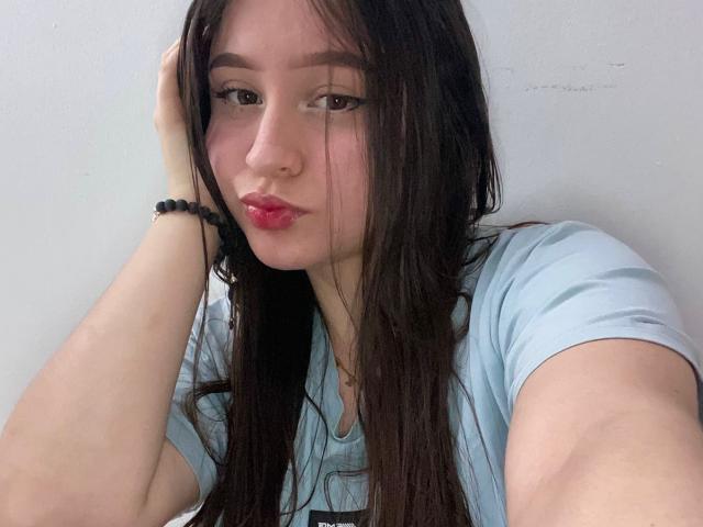 SophieHale with long brown hair, pouty pink lips, wearing a light blue shirt, resting her head on her hand, black bead bracelet on her wrist, slightly messy look.