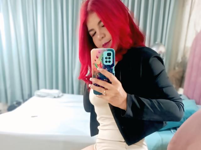 CorineRay with bright red hair, wearing a black jacket over a white dress, taking a selfie in a mirror, showing her small tits, holding a phone with a colorful case.