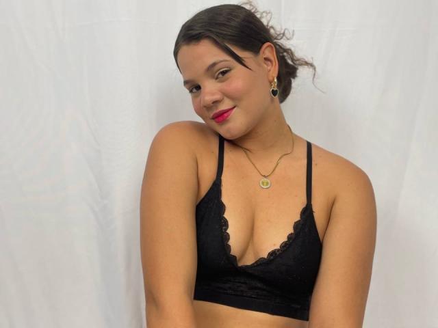LatinXCouple69 con capelli scuri, rossetto rosso, reggiseno di pizzo nero, collana dorata, orecchini e un leggero sorriso, in piedi su uno sfondo bianco, che mostra il suo seno di medie dimensioni..