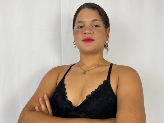 LatinXCouple69 ha seni di media grandezza, indossa un reggiseno di pizzo nero, rossetto rosso e orecchini dorati, con le braccia incrociate e un leggero sorriso, su uno sfondo bianco..
