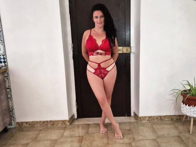 ReifeVenitta-hot steht barfuß in roter Spitzenunterwäsche mit Trägern, zeigt ihre Brüste und Vulva, Hände hinter dem Rücken, langes schwarzes Haar, lächelnd, in einem Türrahmen..