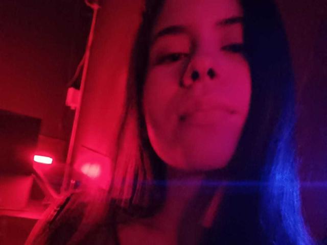 CrystalBrent with blue hair, red-pink lighting, nose ring, and slight pout, looking down, blurry, with a visible lens flare, in a low-light, intimate setting.