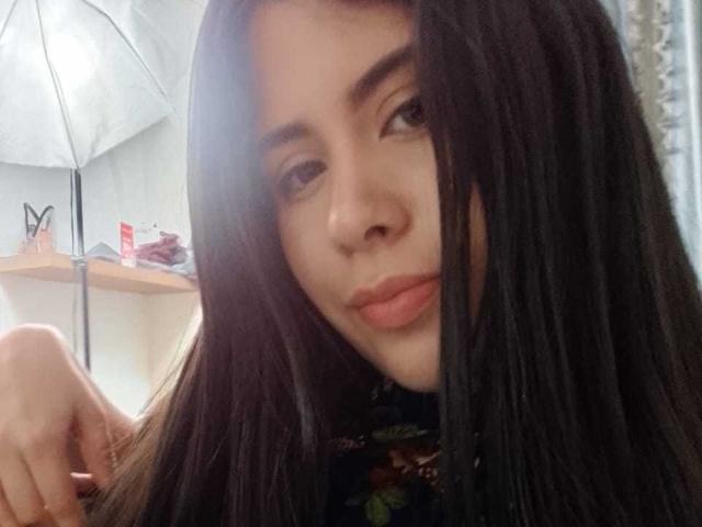 CrystalBrent with long black hair, slightly parted lips, and a subtle smile, holding her hair on the left, wearing a floral top, soft light on her face.