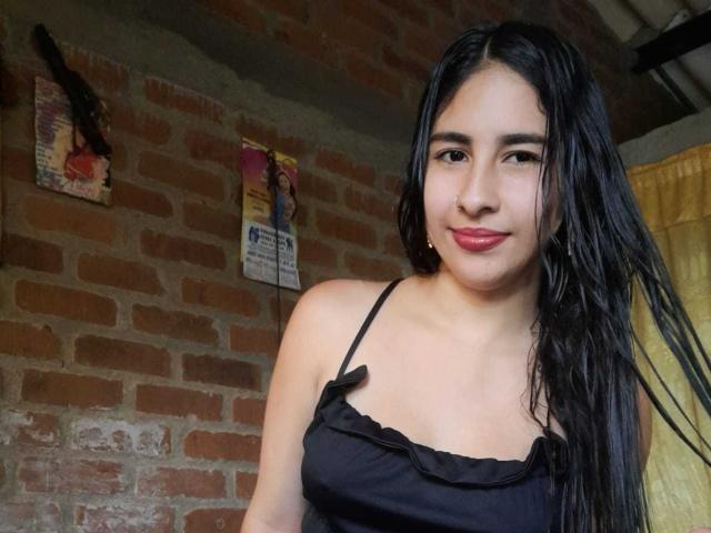Katnis has wet black hair, wearing a black spaghetti-strap top with small bows, showing her tits, smiling with pink lipstick, against a brick wall.