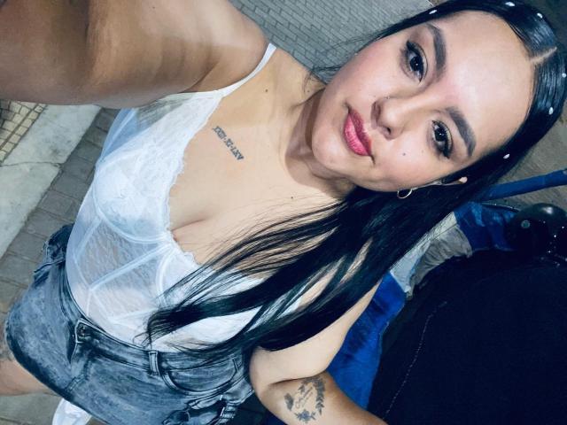 Tindra Zavaleta met lang zwart haar, een witte kanten top die haar borsten laat zien, en grijze denim shorts, lachend naar de camera, een kleine tatoeage boven haar rechterborst, roze lippenstift..
