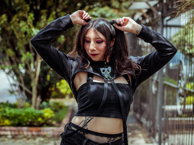 EssledthAnne with dark hair, wearing a black leather crop top and suspenders, has arms raised, showing small tits and a tattoo under her boobs, in a green outdoor setting.