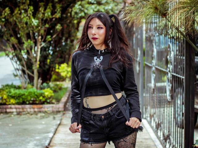 EssledthAnne has dark hair, red lipstick, black leather crop top, high-waisted black shorts, fishnet stockings, and a beige belt. She stands outside, holding straps, looking confident.