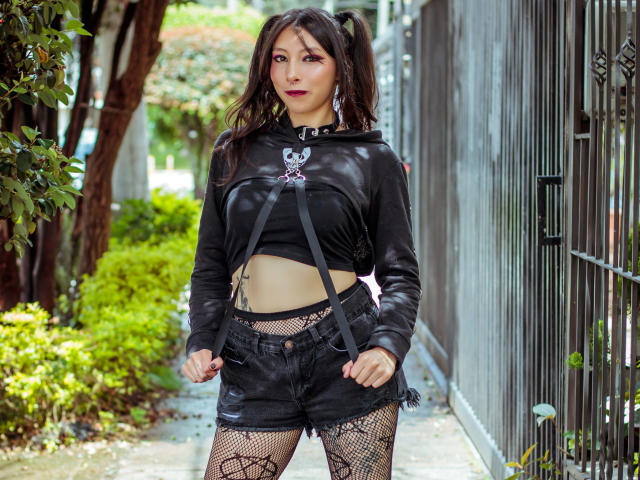 EssledthAnne has black pigtails, wearing a black leather crop top, black shorts, fishnet leggings, and a spiked choker. She's standing with a confident look, hands on her shorts.