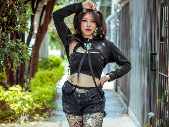 EssledthAnne with pigtails, black leather top, black shorts, fishnet stockings, and suspenders, standing with hand on head, showing tattoos and a small skull necklace. She's got a serious face.