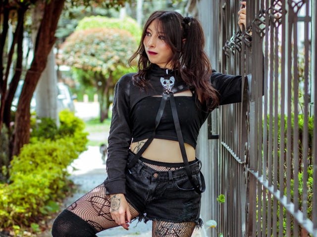 EssledthAnne with dark hair, black top, black shorts, fishnet stockings, and a skull necklace, leaning on a gate, showing tattoos and a confident, sexy look.