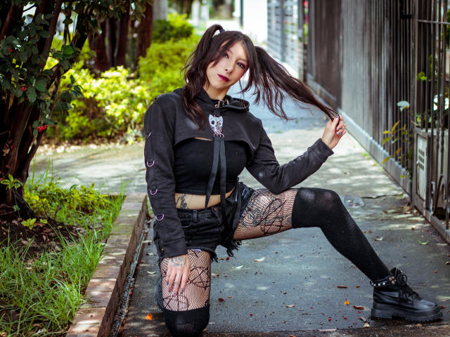 EssledthAnne with dark pigtails, black crop top, and shorts, fishnet legs, knee up, holding hair, black boots, tattoos, and a cute face, in a street setting.