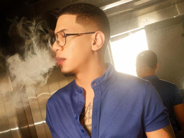 JoshMiiller, com cabelo preto curto, óculos e camisa azul, fumando, com fumaça branca ao redor do rosto, tatuagem visível no peito, reflexo na janela, parte de trás da cabeça no espelho..