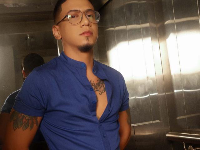 JoshMiiller with glasses, blue button-up shirt, chest tattoo, and tattooed arm, standing in a dimly lit room, looking slightly up, hands in pockets, short dark hair, goatee.