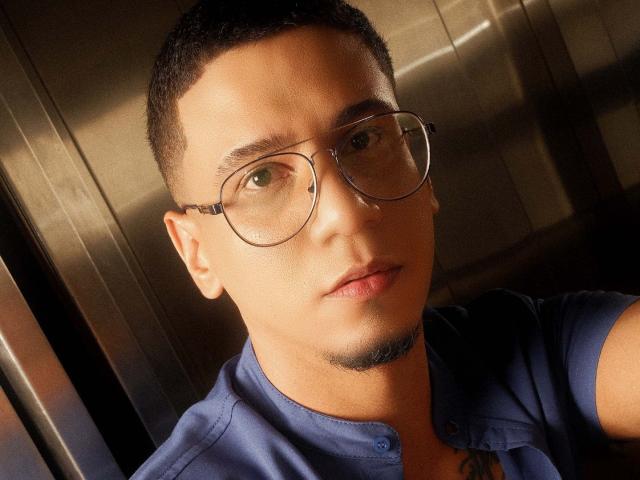 JoshMiiller with short black hair, glasses, blue shirt, and stubble, looking at camera with a slight smile, sunlight highlighting his face. His medium brown skin and tattoos are visible.