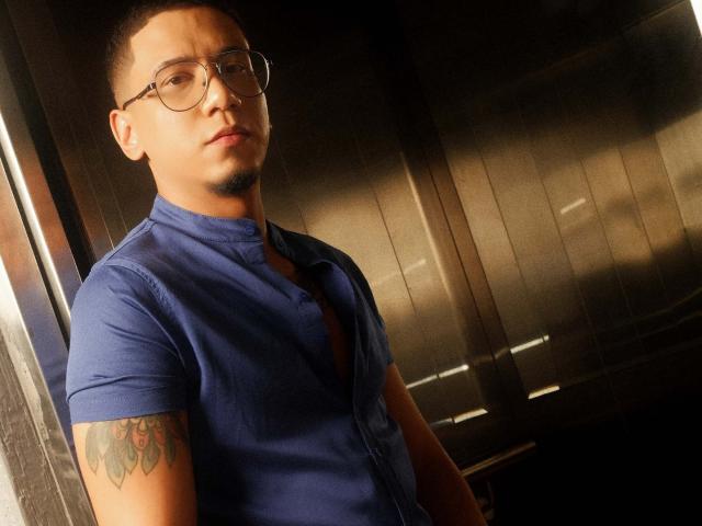 JoshMiiller, de óculos, vestindo uma camisa azul, com um pouco do peito à mostra, tatuagem no braço direito, em pé em uma luz fraca, com uma expressão séria. Fundo de madeira escura..