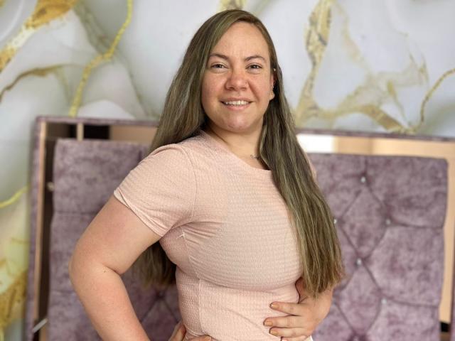 TalianaCediel has long greenish-brown hair, smiling, wearing a pink ribbed shirt, hand on her belly, showing her tits, standing in front of a purple tufted headboard.