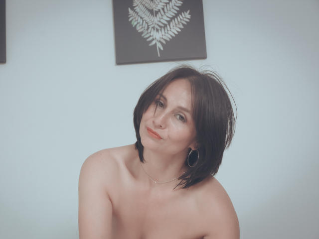 LatinLola with short black hair, small hoop earrings, and red lipstick, topless, smiling slightly, looking at camera, pale skin, subtle makeup, soft lighting, gentle expression, white background.