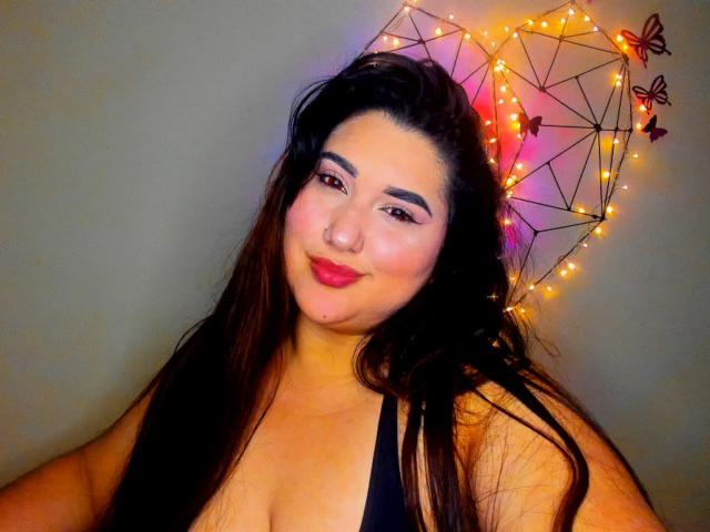 Madeleine Deveraux mit langen, dunklen Haaren, die ein schwarzes Oberteil trägt, lächelnd, mit pinkem Lidschatten, rotem Lippenstift und einer Wanddekoration aus Draht mit orangefarbenen Lichtern und Schmetterlingsmotiven..