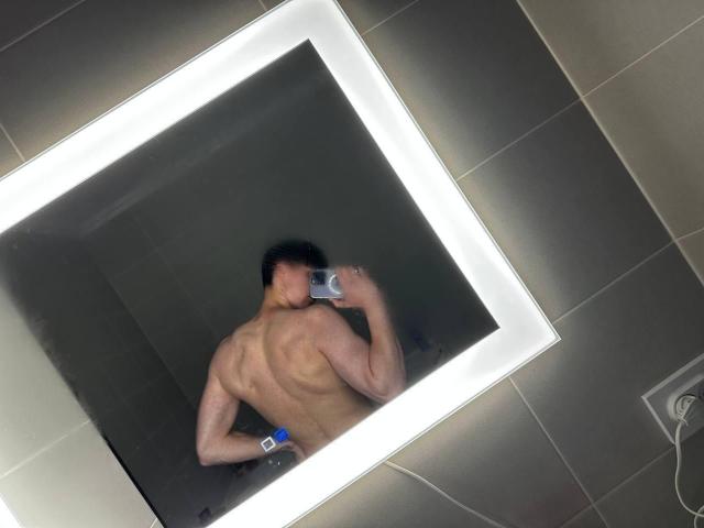 Mirror selfie of VipCouples with a muscular back, holding a phone, showing strong, defined muscles, and a blue wristband. Reflection in a brightly lit, rectangular mirror.