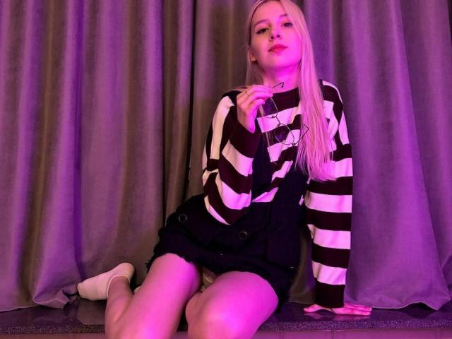 VipCouples hat langes, blondes Haar, ein gestreiftes schwarz-weißes Hemd, schwarze Shorts, weiße Socken, sitzt im violetten Licht, hält ein Glas in der Hand, schaut in die Kamera und lächelt..