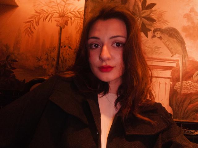 SunnyLeila with red lipstick, wavy brown hair, and dark green jacket, smiling at the camera. Her tits are small, and she has a cute face. Warm, orange lighting.