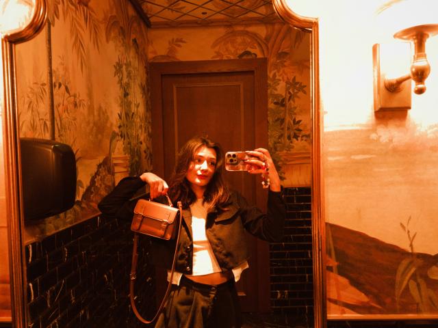 SunnyLeila with long brown hair, wearing a black jacket and white top, holding a brown bag, taking a mirror selfie in a warmly lit, decorative bathroom.