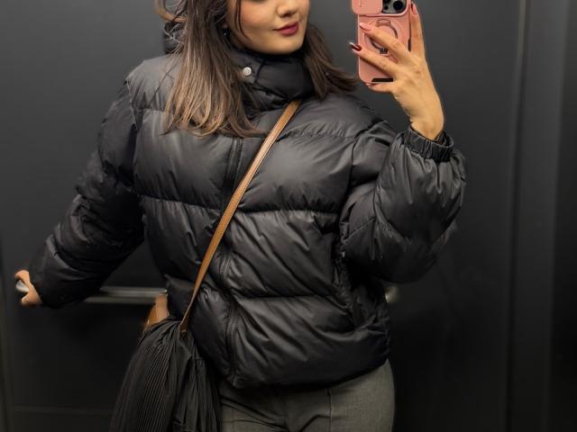SunnyLeila with dark puffy jacket, black shoulder bag, holding pink phone, standing in dark room, wavy brown hair, red lipstick, gray pants, right arm on metal bar.