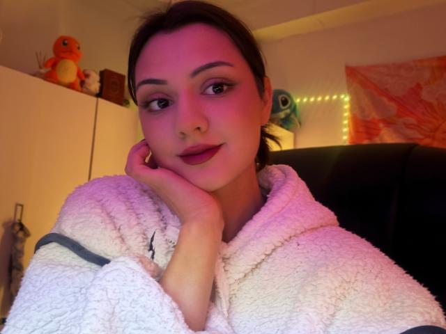SunnyLeila with purple-pink lit face, brown hair, and dark lipstick, wearing a fluffy white hoodie, resting her chin on her hand, looking at the camera with a slight smile.