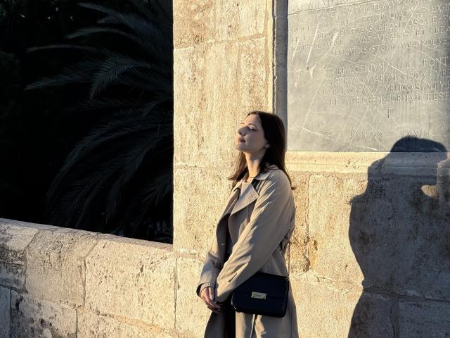 SunnyLeila stands in sunlight, shadow on wall, wearing beige coat, black purse, looking up, brown hair, fair skin, hands near waist, palm leaves in dark background.