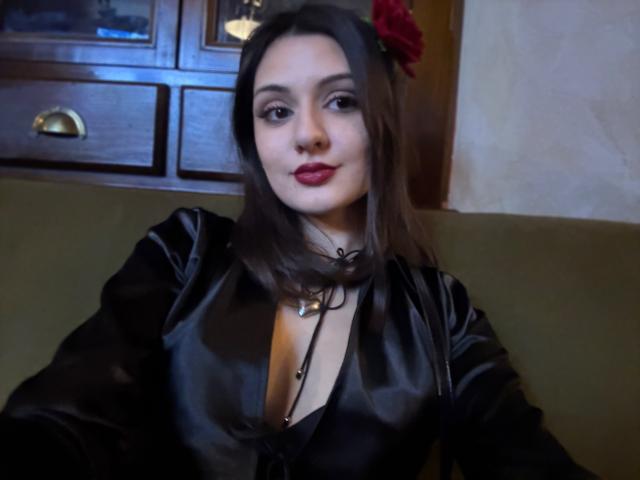 SunnyLeila with dark hair, red rose in hair, black satin dress, low-cut showing small tits, black choker, red lipstick, sitting, wooden cabinet in background, selfie.