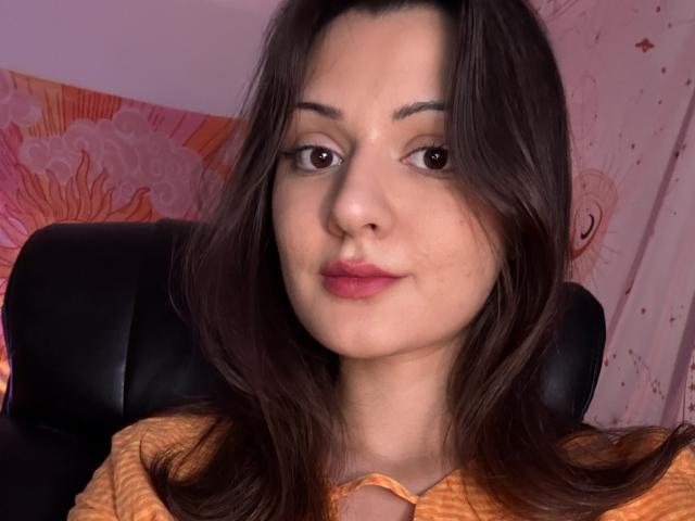 SunnyLeila with long brown hair, brown eyes, and pink lipstick, wearing an orange top, sitting in a black leather chair. Her face has a slight smile.