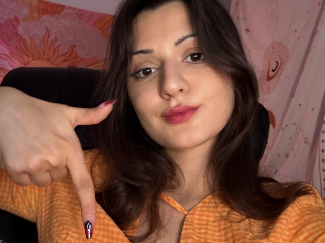 SunnyLeila with brown hair, orange checkered shirt, and pink lips, pointing with her middle finger, showing a small, colorful tattoo on her nail.