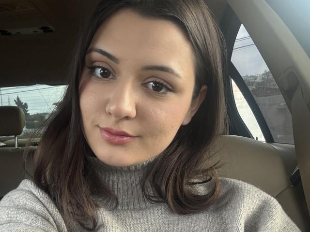 SunnyLeila has a cute face with brown eyes, dark brown hair, and pink lips. She's wearing a grey turtleneck, sitting in a car, looking at the camera.