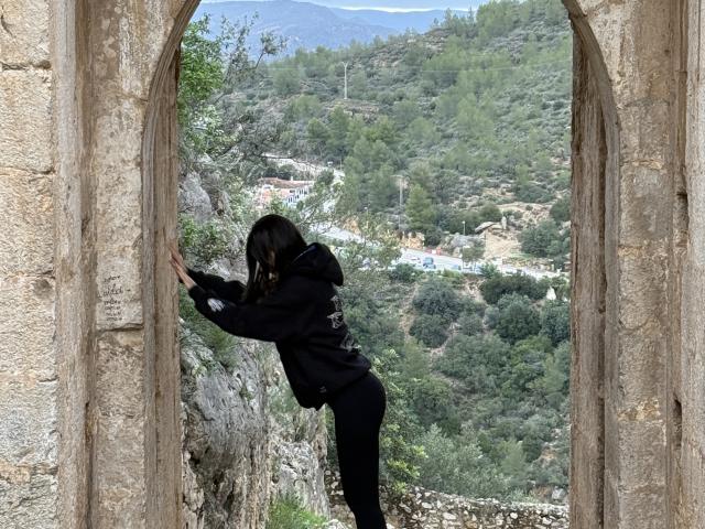 SunnyLeila with black hair, wearing a black hoodie and black pants, leans through a stone archway, looking outside at a green, forested hill. Her ass is slightly visible.