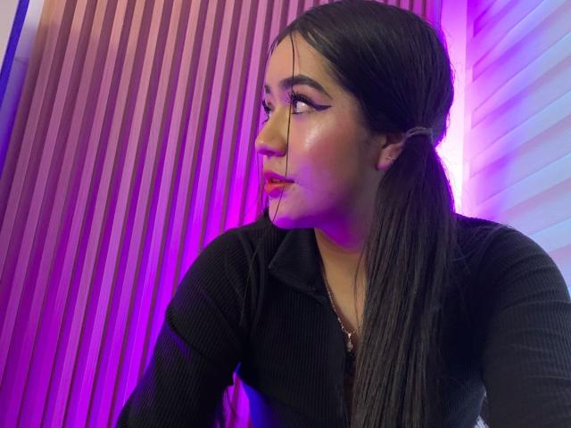 JennaStone with long black pigtails, wearing a black ribbed shirt, has pink and purple neon lighting on her face, looking to the side with glossy pink lips.
