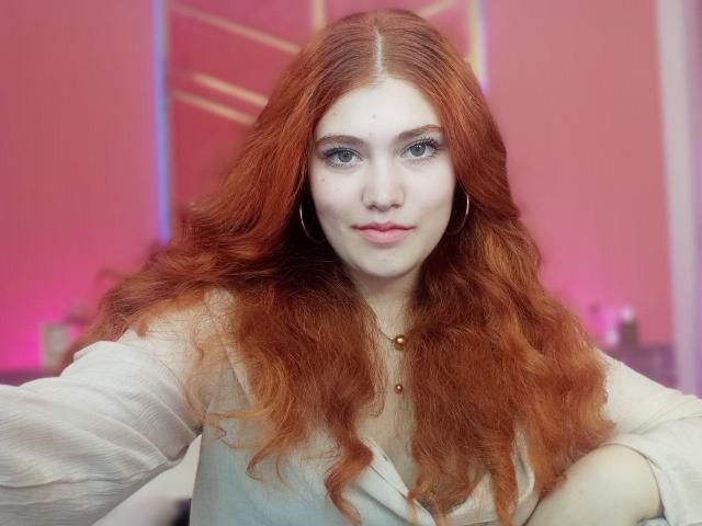 LindaSauver has long red wavy hair, pale skin, blue eyes, and wears a white shirt with gold hoop earrings and a small gold necklace, looking straight at the camera.