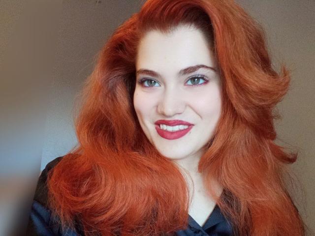 LindaSauver has red, wavy hair, blue eyes, and red lipstick. She's smiling with her tits partially covered by a dark shirt. Her skin is pale and she looks hot.