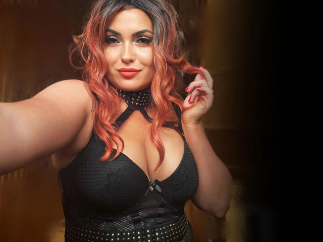MystischLea69-hot with red ombre hair, black cleavage-cut top, and studded collar, holding her hair, showing her tits, and smiling with red lipstick.