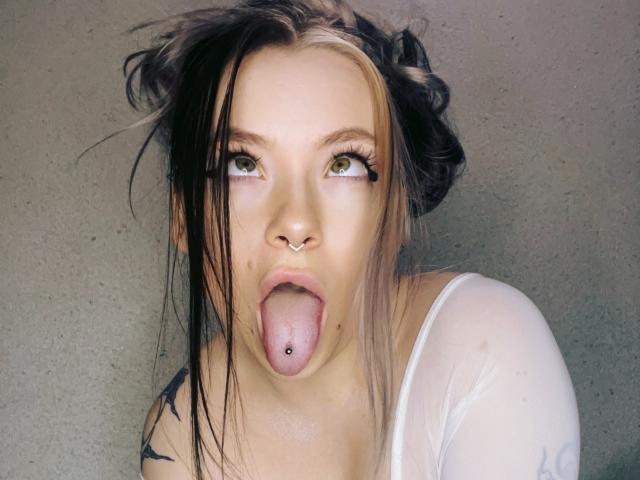 SadPotato with messy black and blonde hair, green eyes, nose septum piercing, tongue pierced, white shirt, looking up with mouth open, showing a bit of a tattoo on the shoulder.
