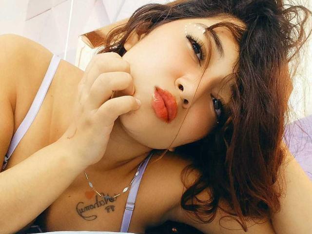 NicoleMartins with dark wavy hair, pouty red lips, wearing a purple bra, hand near mouth, tattoo under chin, shiny eyebrows, soft light, intimate selfie.