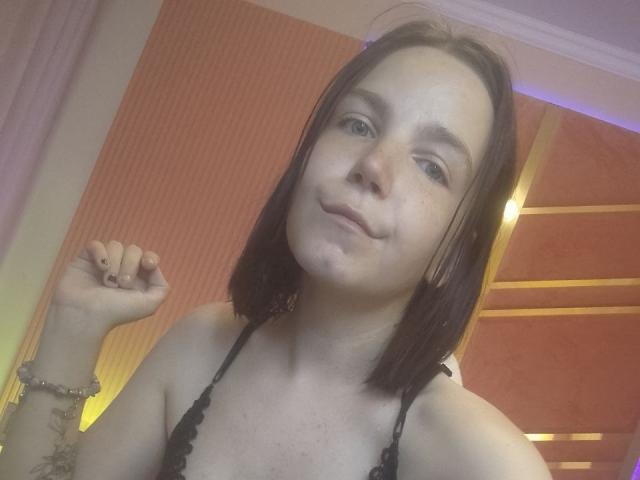 SpicyDora has medium-length brown hair, light skin, and small tits. She's wearing a black lace bra, smiling slightly, with a raised fist, and a bracelet on her left wrist.