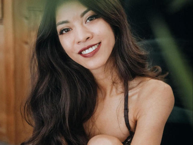 Kim-Rose with long dark hair, smiling with red lipstick, wearing a black strap dress, showing her small tits and cute face. Her brown eyes are looking at the camera.