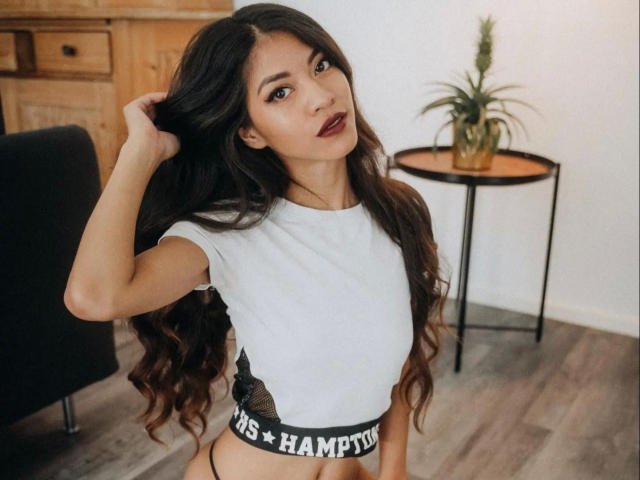 Kim-Rose with long, wavy dark hair, wearing a white crop top and black "HAMPUS" underwear, touching her hair, looking at the camera with dark red lipstick.