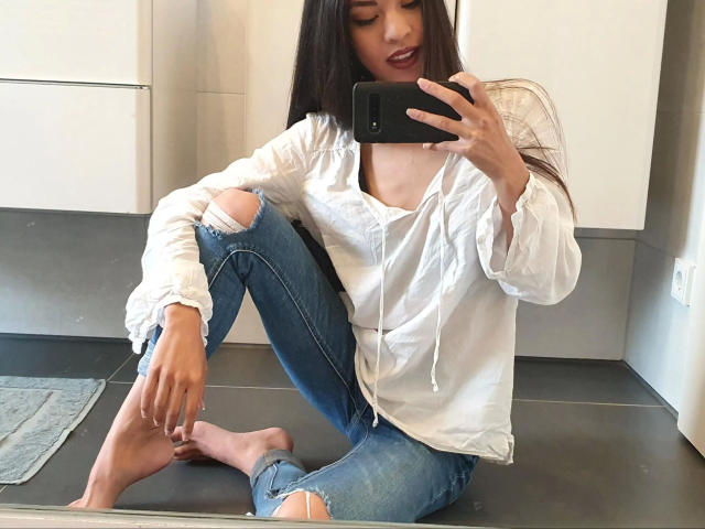 Kim-Rose with long black hair, wearing a loose white shirt and ripped blue jeans, sits barefoot on a kitchen floor, taking a mirror selfie with her phone.