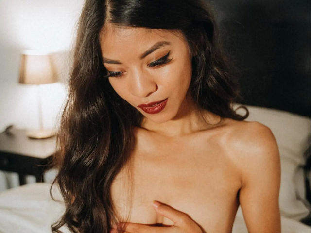 Kim-Rose, topless, with long dark hair, looking down, covering her tits with one hand, brown eyes, winged eyeliner, red lipstick, soft light, bedroom, gentle expression.