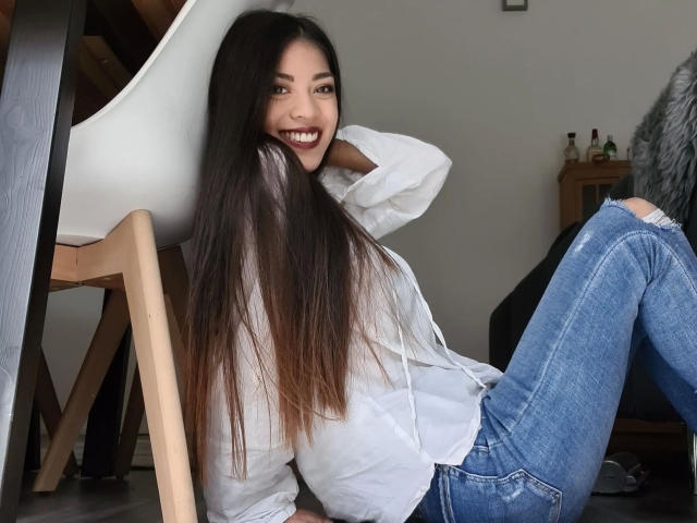 Kim-Rose with long brown hair, white button-up shirt, and blue jeans, smiling with teeth visible, sitting on wooden chair, hand behind head, legs crossed.